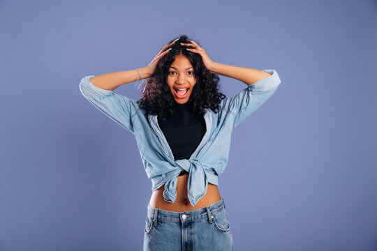 Young woman with curly hair wearing a denim shirt and black top poses with hands on her head against a solid purple background, expressing excitement and joy