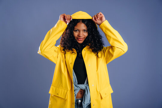 Young woman with curly hair wearing a bright yellow raincoat adjusts the hood while posing against a solid purple background in a studio setting