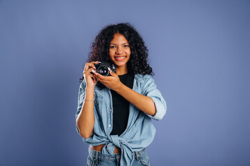 Fototapeta premium Young woman with curly hair wearing denim shirt holds a camera and smiles against a solid blue background, showcasing her interest in photography and creative expression