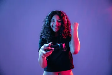 Young woman with curly hair wearing headphones holds a video game controller, celebrating victory against a colorful background with purple and blue lighting © standret