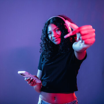 Young African American woman wearing headphones holds smartphone and points finger towards the camera against a colorful gradient background with soft lighting