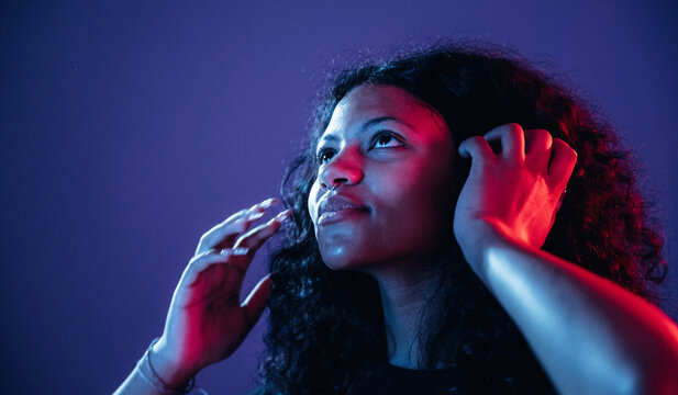 Young African American woman with curly hair adjusts headphones while looking up against a colorful gradient background with purple and red lighting