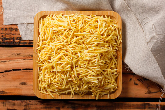 Crispy yellow shoestring potatoes stacked on a square wooden serving platter with a linen cloth in the background
