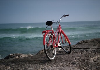 Fototapeta premium Bright red vintage bicycle parked on a rocky stone ledge overlooking the calm blue ocean waves