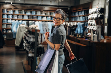 Fototapeta premium Senior woman with short gray hair and glasses smiles while holding shopping bags inside a clothing store with denim items displayed on shelves in the background