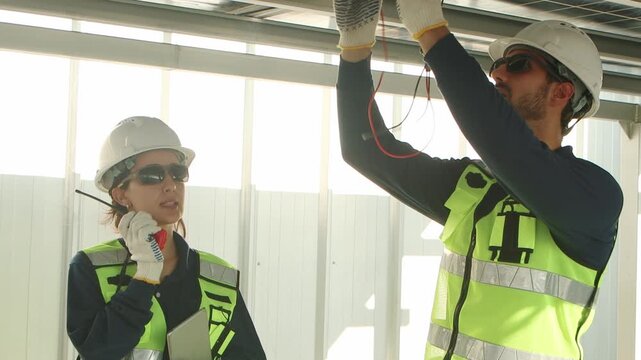 Two engineers with safety vest and helmet walking together to inspect solar panels. Technicians discussing, Man using multimeters to check, woman with tablet speaking into walkie talkie at solar farm.