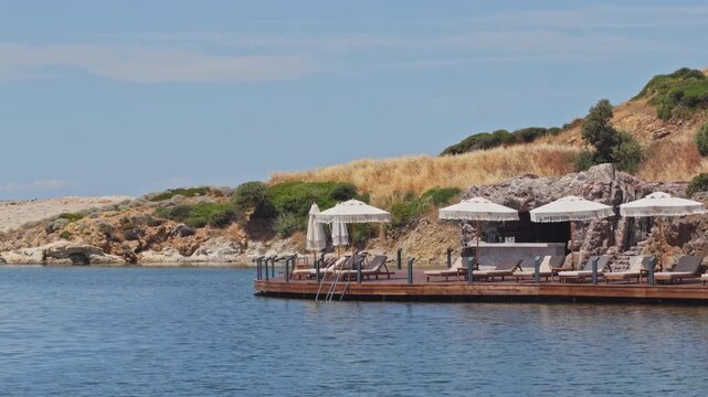 High-quality 4K wide shot of an elegant wooden sun deck over the calm sea at a luxury beach resort. Features white umbrellas, loungers, and a seaside bar.