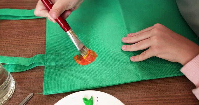 girl painting orange tangerine for capybara on green reusable eco tote bag at home. Creative young artist upcycling fabric shopper with cute custom animal food illustration. Teenager practicing