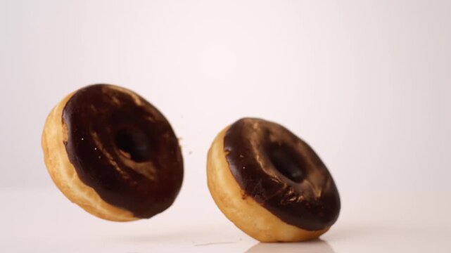 Chocolate-glazed glossy donuts bouncing and tumbling on white in slow motion macro studio shot. donut shop ads, coffee chain commercials, weekend treat reels.