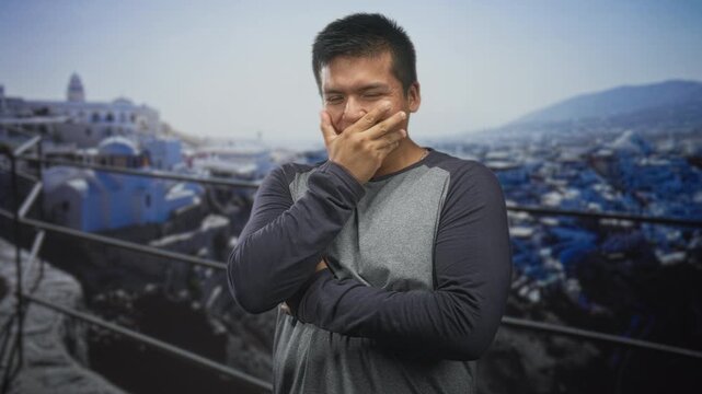 Man with hand over mouth and crossed arms standing on a building balcony overlooking a coastal town and railings, appearing to stifle a laugh; mirth.