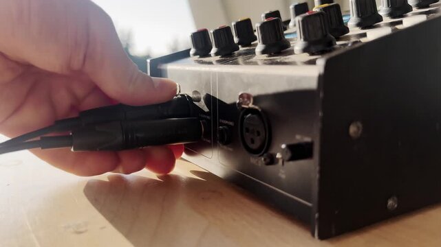 Close-up of a hand unplugging a cable from a broadcast intercom audio unit, with control knobs, headset socket, sunlight, and live production equipment detail.