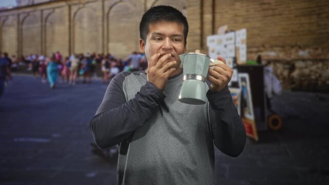 Young man holding a coffee maker by its handle, fingers to mouth in a crowded street with stone wall and market stalls; surprise curiosity.