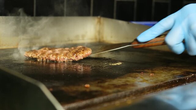 Close-up view of a cook in blue gloves using a press to smash and fry a fresh ground meat patty on a hot, steamy restaurant griddle, preparing a delicious burger for a customer