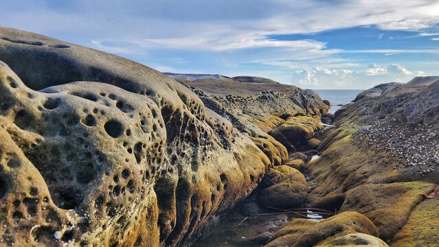 Panoramic landscape of massive coastal rock formations with distinct honeycomb patterns lining a narrow channel leading toward the sea.