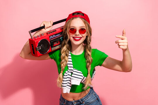 Young hipster girl with blonde braids green shirt red cap poses with a red boombox smiling and pointing against pink background