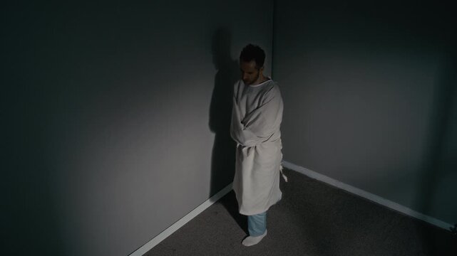 High angle of Caucasian adult man wearing straitjacket pacing in dark corner of psychiatric ward cell. Footage is suitable for mental health awareness campaigns or medical documentaries