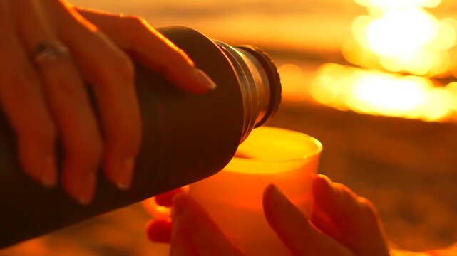 Pouring tea thermos hands filling a cup on the beach during a peaceful golden hour sunset over the sea