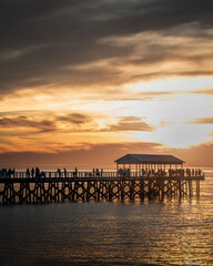 Fototapeta premium People gathered on a wooden pier at sunset with dramatic clouds over calm ocean waters in South Australia