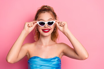 Naklejka premium Smiling young woman with braces wearing white cat-eye sunglasses and blue strapless dress against pink background