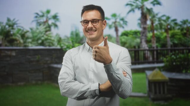 Man with moustache and glasses giving thumbs up with right hand in a building courtyard, striped shirt visible; confidence.