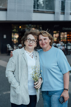 Two Senior Female Friends With Chamomile Flowers Near Modern Housing