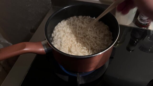 Horizontal 16:9 4K video of instant noodles beginning to boil inside a pot on a kitchen stove, with chopsticks stirring the soup.