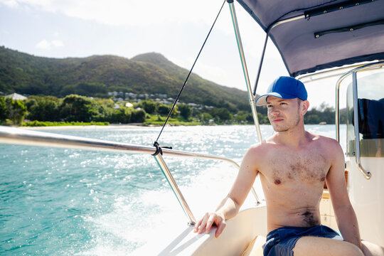 Shirtless male tourist with cap sitting in speedboat cruising along tropical coastline of Praslin island in Seychelles. Summer vacation, ocean travel, luxury lifestyle and island paradise concept.