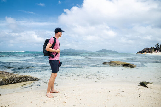 Male tourist with backpack and cap standing on tropical beach in Seychelles, looking around at scenic landscape. Summer vacation, island exploration, freedom and solo travel concept.
