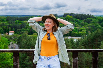Obraz premium Smiling woman in hat standing on observation deck against green forest valley background. Vacation, tourism and travel success concept.