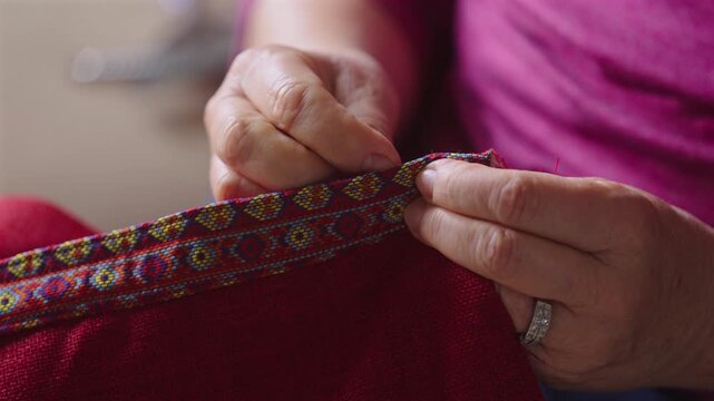 Elderly ringed hands needle red thread through a colorful folk-patterned woven trim
