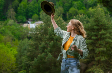 Profile portrait of smiling woman raising hat against green forest background. Joyful mood and freedom concept. © 5ph