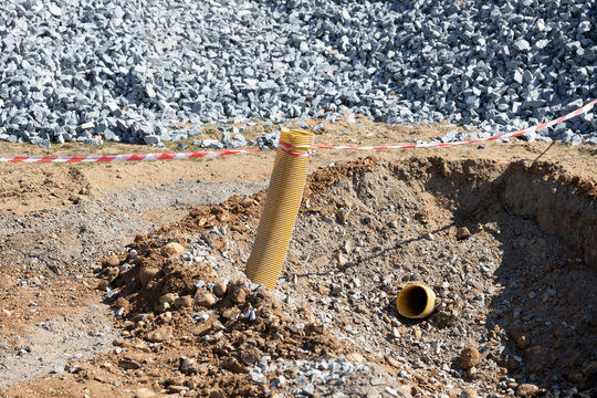 Yellow drainage pipe protruding from a construction site trench surrounded by gravel and soil, with safety tape marking the area for caution