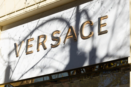 Storefront sign displaying the logo "VERSACE" in gold lettering against a white background with shadows from nearby trees in a city setting