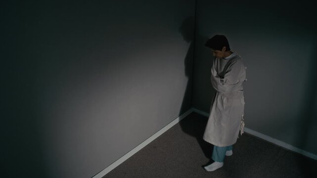 High angle shot of mature Caucasian female patient wearing straitjacket pacing in dark psychiatric clinic room. Footage is ideal for mental health awareness campaigns or medical documentaries