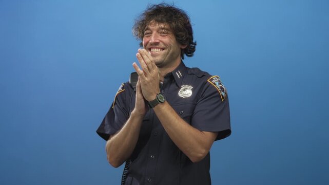 Man in police uniform smiling and clapping hands with badge and radio visible against blue backdrop in studio; pride duty.