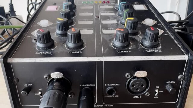 Close-up of a broadcast intercom control unit with comm knobs, sidetone dials, XLR microphone inputs, headphone jacks, and connected production cables.