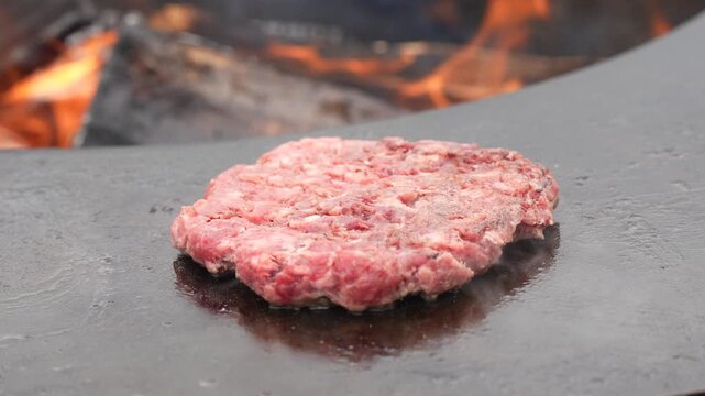 Fresh raw minced beef burger patty sizzling on a hot circular steel plancha over burning wood fire for outdoor backyard barbecue catering concept