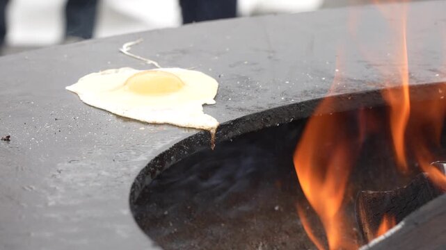 Single fried egg with a bright yellow yolk sizzling on a hot circular black plancha griddle over a burning wood fire during an outdoor culinary event