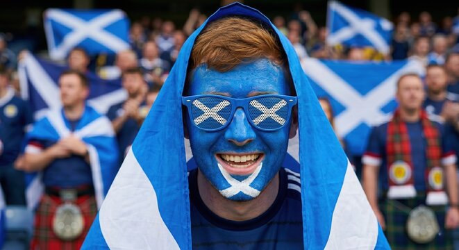 Portrait of a cheerful Scotland soccer fan with face paint and novelty glasses, draped in the national flag. Happy supporter smiling at a stadium during an international sports championship.