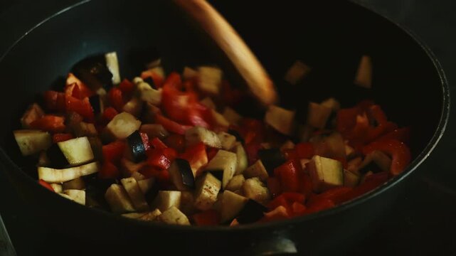 Cooking vegetables, preparing plant based vegetarian meal. Hand with a wooden spoon stirs red bell peppers and eggplant in frying pan