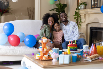Obraz premium Parents and son sitting on white couch in living room with rainbow cake and balloons