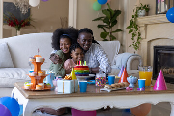Obraz premium African American family sitting in living room, hugging child, blowing lit candles on rainbow cake