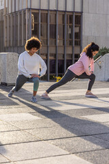 Two adult women stretching and lunging on sunlit concrete plaza near office building windows © wavebreak3