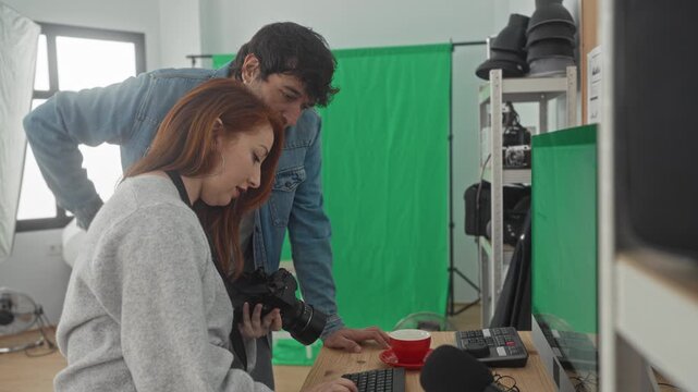 Woman holds camera and leans toward a desktop computer while both photographers study images on the monitor in a studio; creative focus.