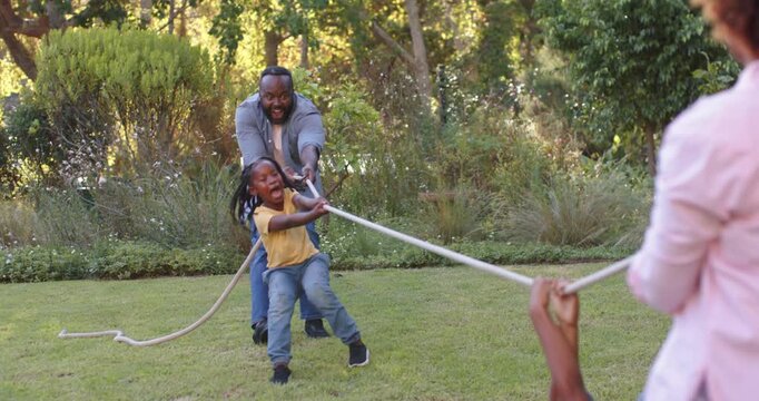 African family playing rope tug on lawn, dad guiding boy in yellow shirt while opponents pulling