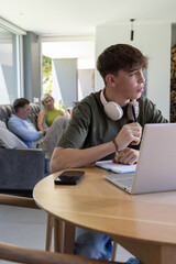 Obraz premium Family with teenage son sitting at round wooden table, studying on laptop with headphones