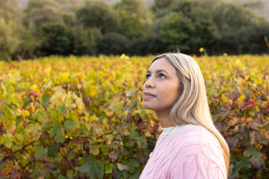 Woman standing in vineyard wearing pink cable-knit sweater, pendant necklace looking up, copy space