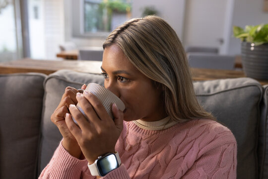 African American woman holding and sipping mug while wearing sweater and smartwatch in living room