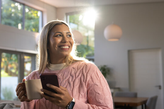 Mid-adult African woman wearing pink sweater standing at home holding phone and ceramic mug