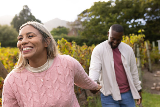 African American couple walking hand in hand through grapevines on path, woman wearing pink sweater